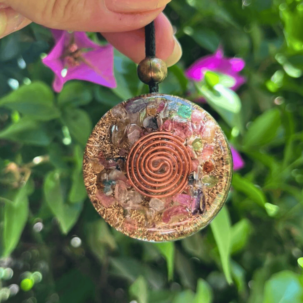 Amuleto de resina con cristales y espiral de cobre, colgado en plantas verdes y flores moradas.